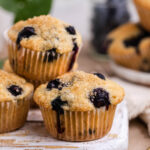 Blueberry muffins on a white serving board ready to eat. With fresh blueberries and coarse sugar on a gold spoon on the side.