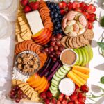 Kid's charcuterie board on pink marble table with plates, a cheese knife, beige napkin, trailing plant, and two glasses of soda.