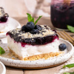 A slice of blueberry dessert topped with fresh blueberries, cookie crumbs, and mint leaves on a white plate with a fork and extra blueberry filling in the background.