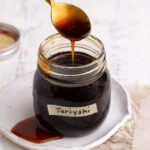 Spooning out a spoonful of teriyaki sauce from a glass jar with a cloth on the side.