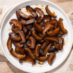 Overhead shot of simmered shiitake mushrooms on a white plate with beige cloth underneath.
