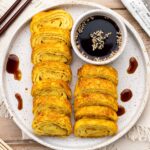 Flatlay shot of vegan tamagoyaki on a white speckled plate with soy sauce, chopsticks, and beige cloth.