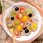 Fruit jelly in a white bowl with a spoon on top of a pink cloth on a beige wood background.