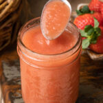 Strawberry applesauce in a glass jar accompanied by a spoon coming out.