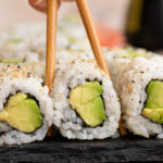 Chopsticks are going in to grab one piece of avocado roll sushi.